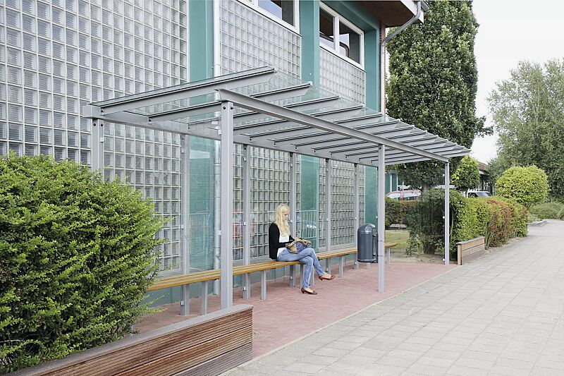 wartehalle-pegasus__shoKAT0029885 Wartehalle PEGASUS, Dachbreite x Dachtiefe 9300 mm x 2000 mm, Rück- / Seitenwände ESG, Klarglas, Sitzbank FERMO und Abfallbehälter DERBY, Stahlkonstruktion in RAL 9006 weißaluminium